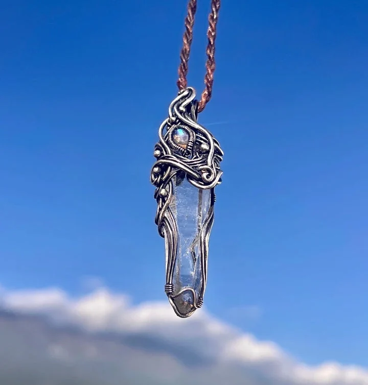 A silver pendant featuring a clear quartz crystal, designed with intricate wire wrapping and a small blue gemstone at the top, hanging against a blue sky with clouds.