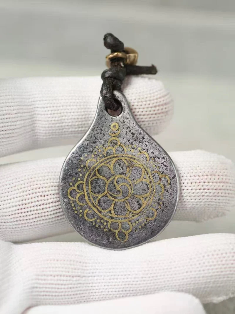 A teardrop-shaped pendant with a distressed silver finish and gold-toned mandala-style detailing, hanging from a black cord, held by white textured gloves.