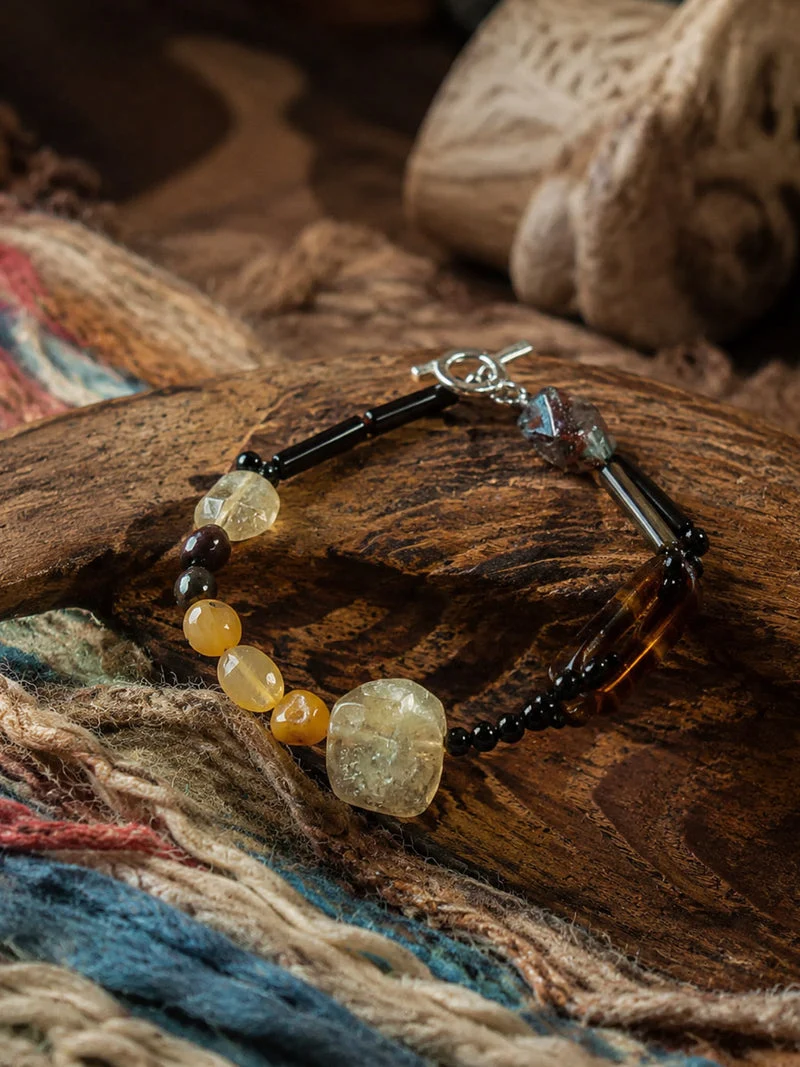 A beaded bracelet with black beads, amber-yellow beads, and a large faceted clear crystal pendant, displayed on a rustic wooden surface with a colorful woven textile underneath.