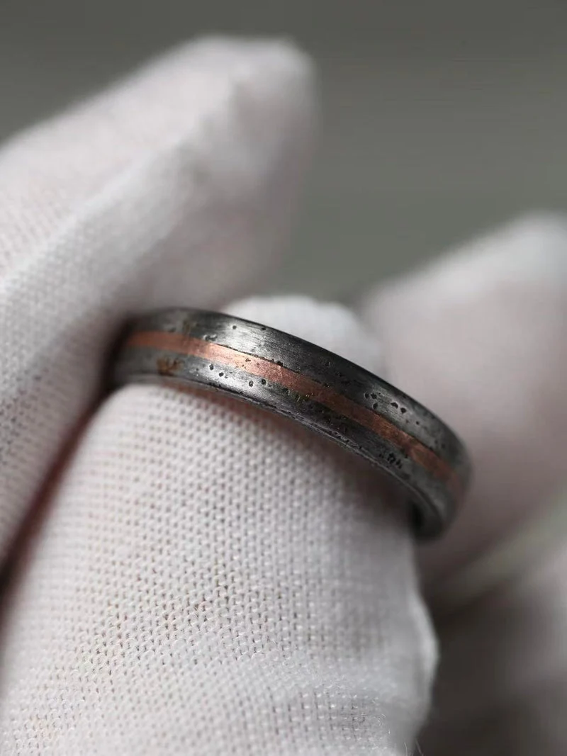 A close-up of a textured silver ring with a central copper inlay, held on a white fabric-covered finger.