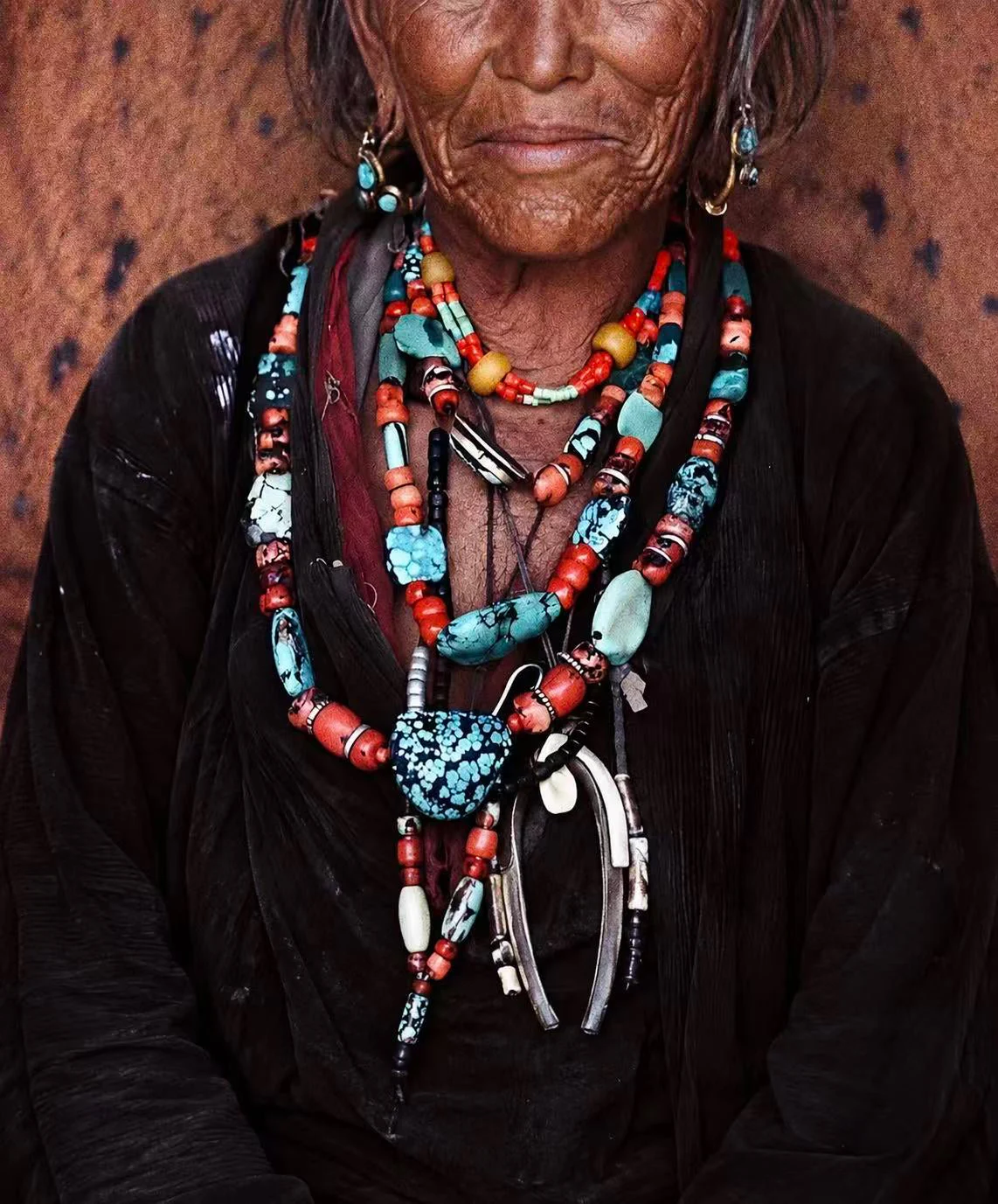 A close-up portrait of an elderly woman wearing multiple layered beaded necklaces and earrings. The jewelry features turquoise, coral-red, white, and black beads, with a prominent turquoise and black patterned pendant.