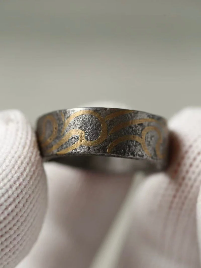 A close-up of a textured silver ring with gold engraved swirl patterns, held by white gloved hands against a soft gray background.