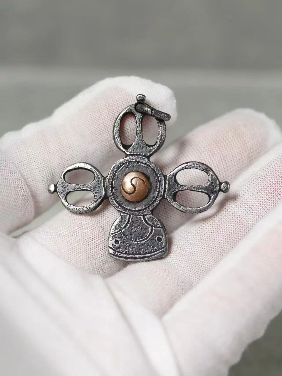 A small ornate pendant or charm with a silver-toned metal design and a central gold-toned spiral detail, displayed on a white gloved hand against a soft gray background.