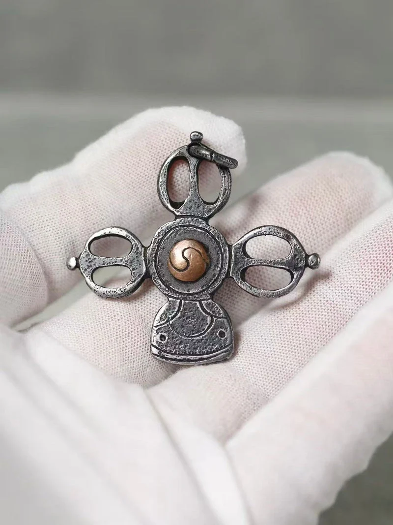 A small ornate pendant or charm with a silver-toned metal design and a central gold-toned spiral detail, displayed on a white gloved hand against a soft gray background.