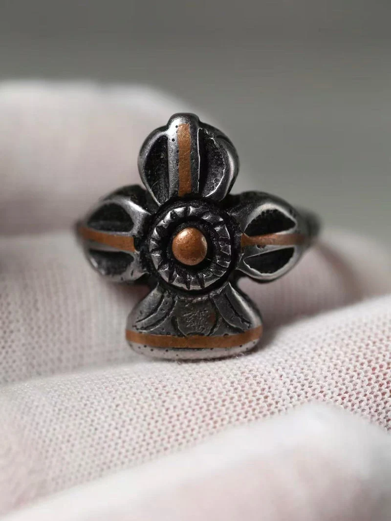 A silver-toned metal ring with a decorative cross-like floral design and a small round gold-toned center, resting on a textured white fabric surface.