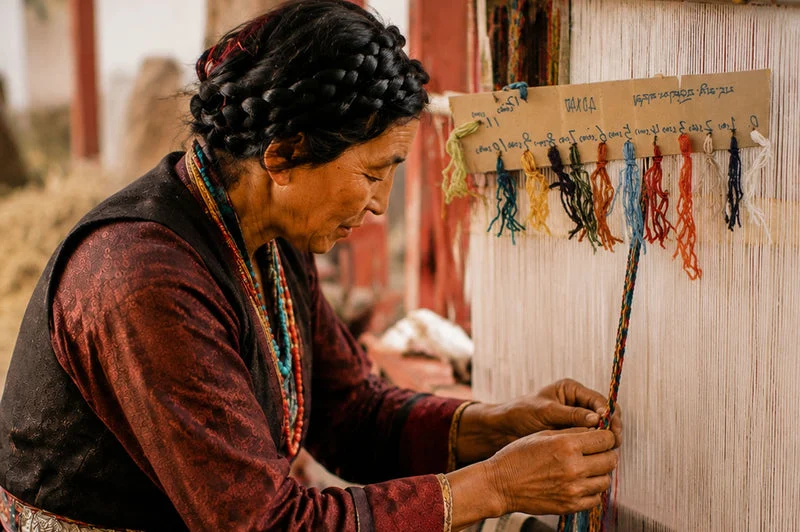 A woman in traditional clothing works on a handloom, weaving a long, narrow textile with colorful thread tassels hanging on a wooden board behind her.