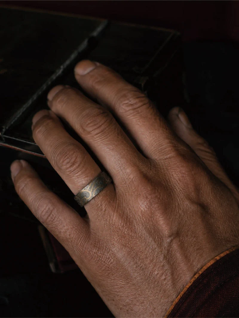 A close-up of a person's hand resting on a dark surface, wearing a simple metallic band ring on the ring finger.