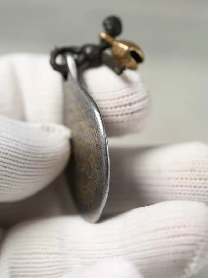 A close-up of a hand wearing white knit gloves holding a small oval metal pendant or charm with a dark loop and a gold-toned top detail.